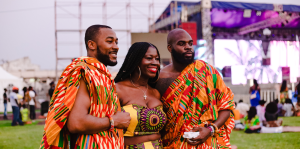Three individuals in vibrant clothing stand together, smiling at an outdoor event celebrating cultural diversity.